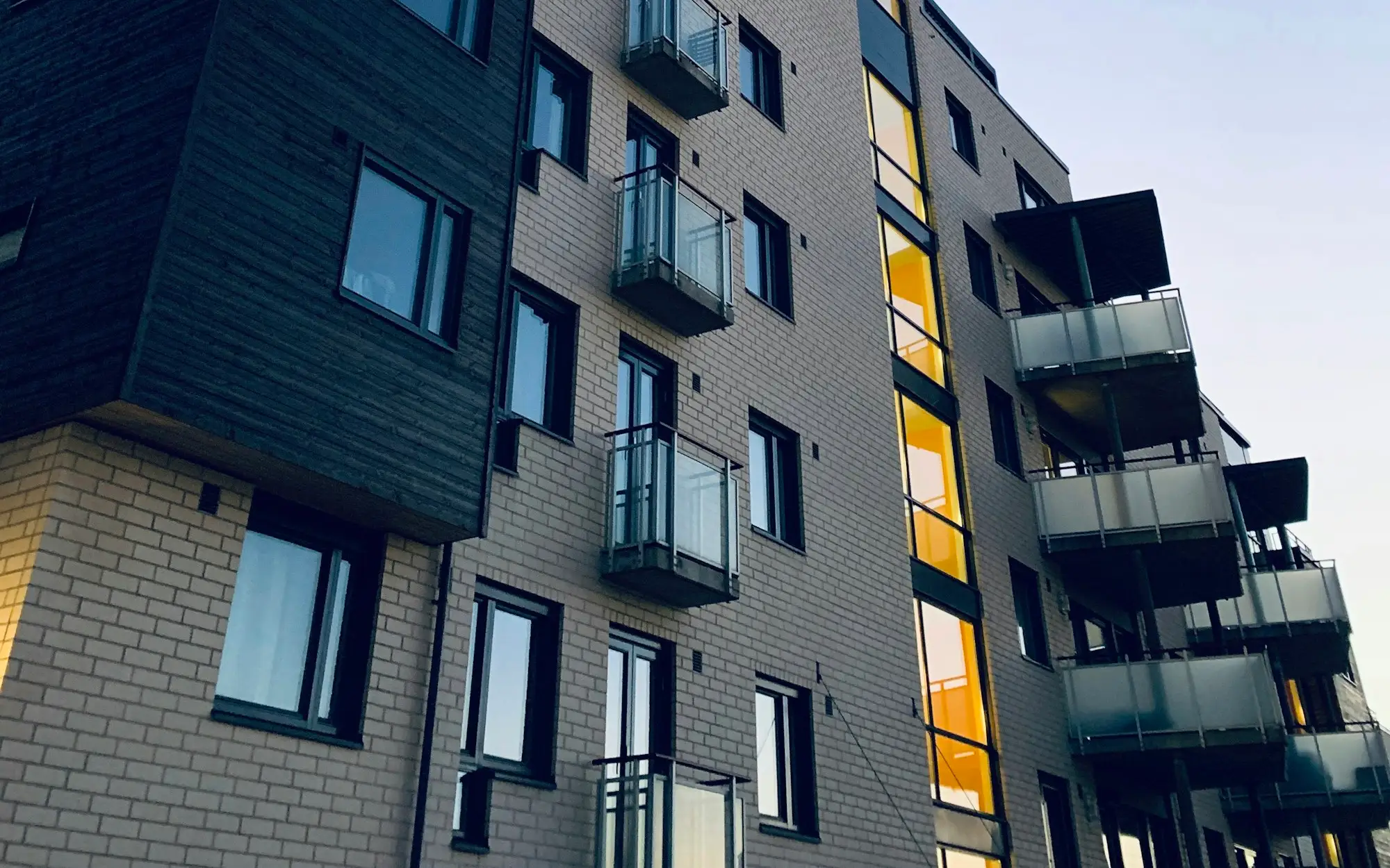 Fachada de un edificio residencial nuevo, con revestimiento de madera oscura, ladrillo claro y balcones, bajo un cielo despejado
